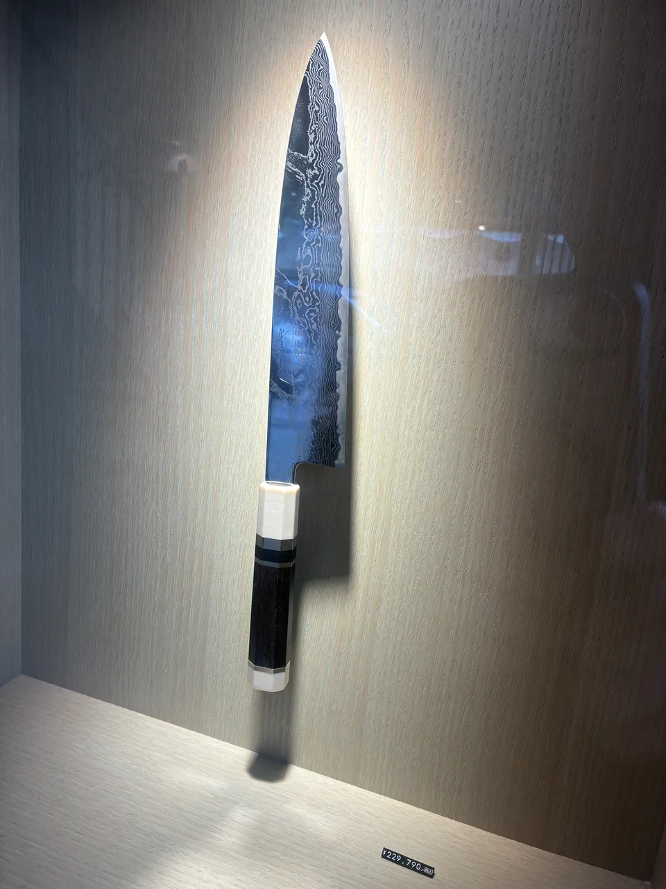 A single damascus steel Japanese kitchen knife with a black and white handle displayed horizontally inside a glass case at a Kappabashi knife shop