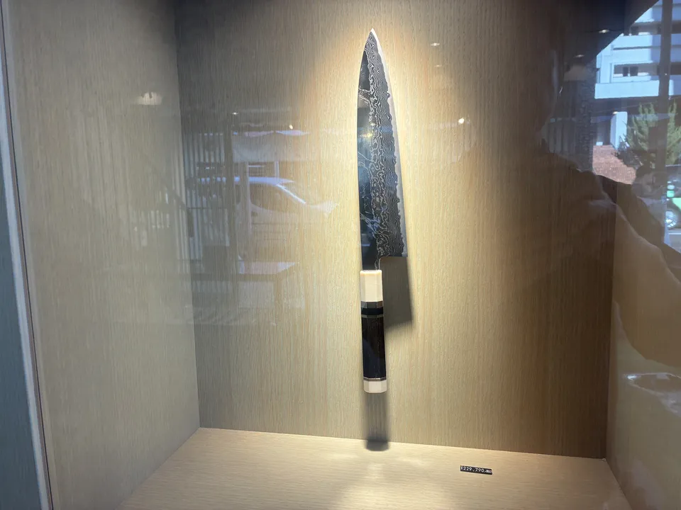 A single damascus steel Japanese kitchen knife with a dark handle displayed vertically in a backlit wooden display case at a Kappabashi knife shop