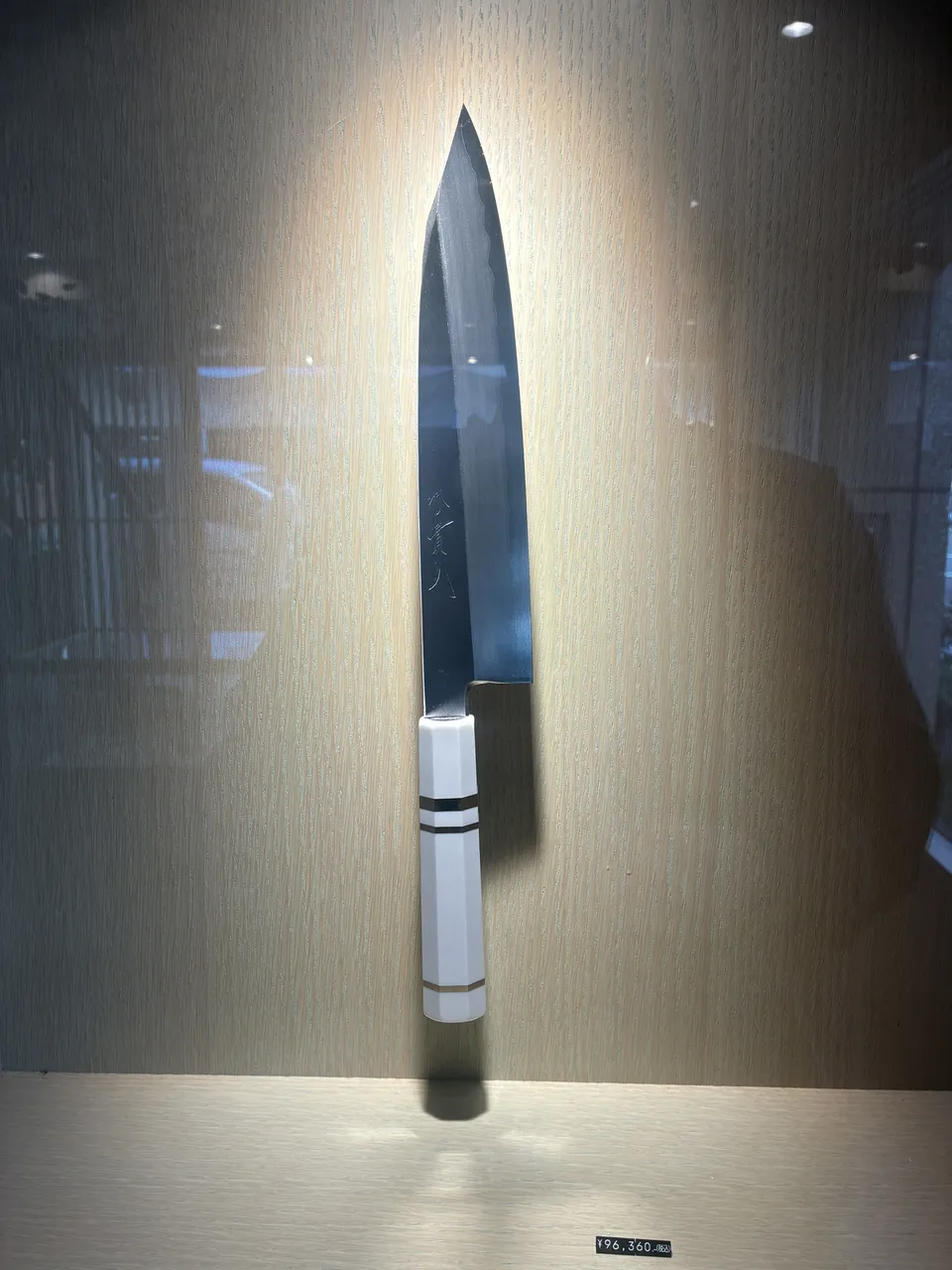 A Japanese kitchen knife with a white handle banded in black rings displayed horizontally inside a glass case at a Kappabashi knife shop