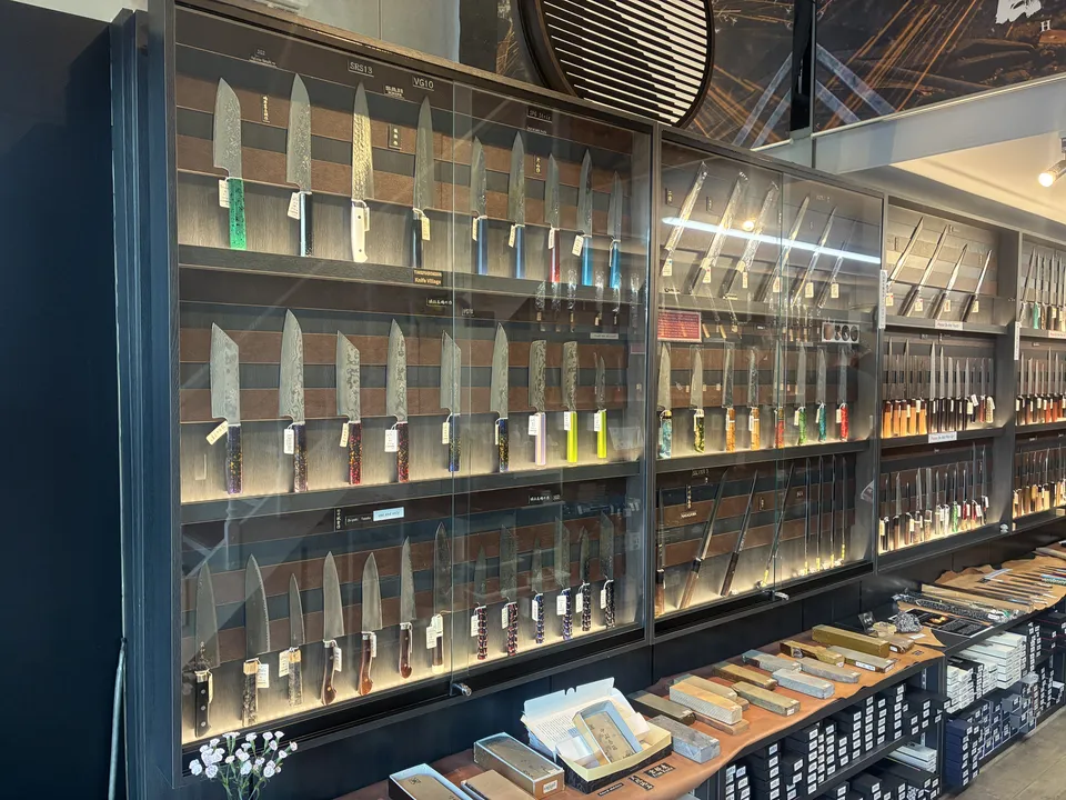 A floor-to-ceiling glass wall display of Japanese kitchen knives with colorful handles, with whetstone boxes arranged on the counter below at a Kappabashi knife