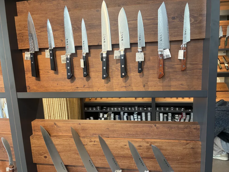 Japanese kitchen knives arranged on a two-tier wooden display rack at a Kappabashi knife shop, with varied blade shapes and mixed black and wooden handles