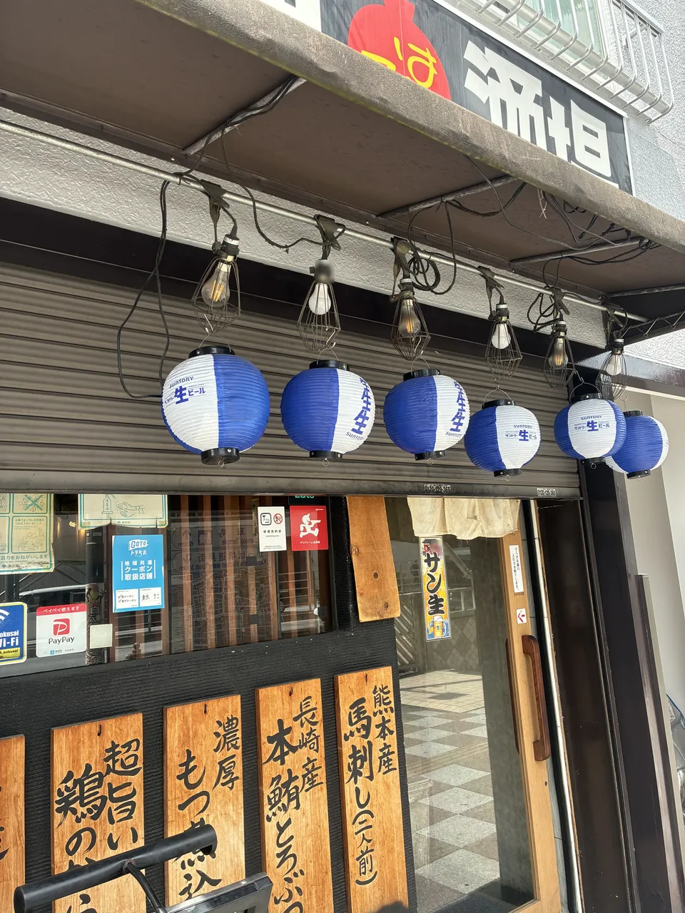 Five blue and white round paper lanterns with Japanese text hanging from the eave above an izakaya entrance in Asakusa, with wooden menu boards and a wooden doo