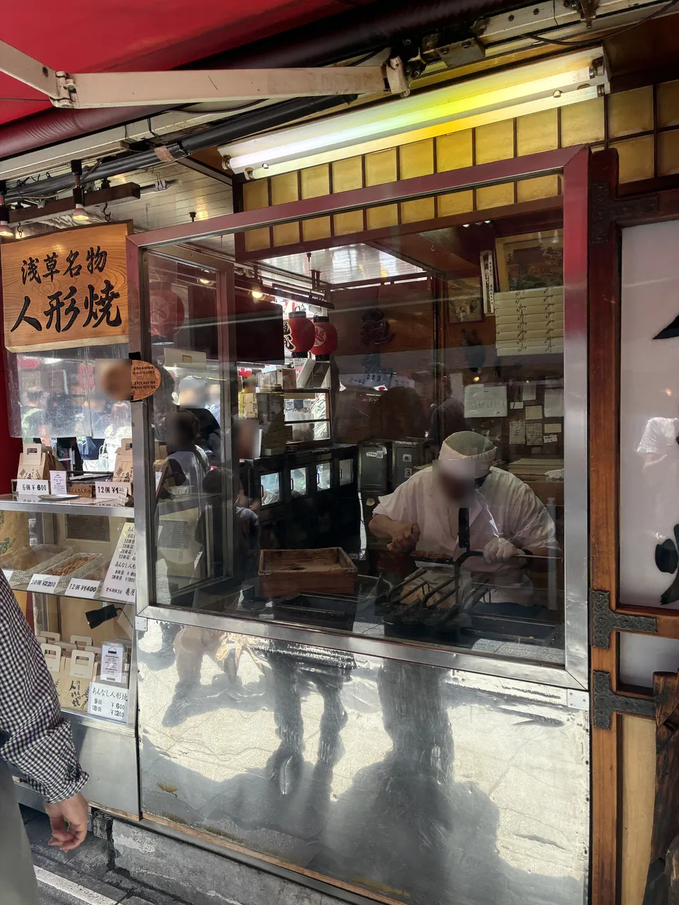 Rotated view through the glass window of a 浅草名物 人形焼 (Asakusa specialty ningyo-yaki) shop on Nakamise, showing a baker in white uniform filling cast-iron molds a