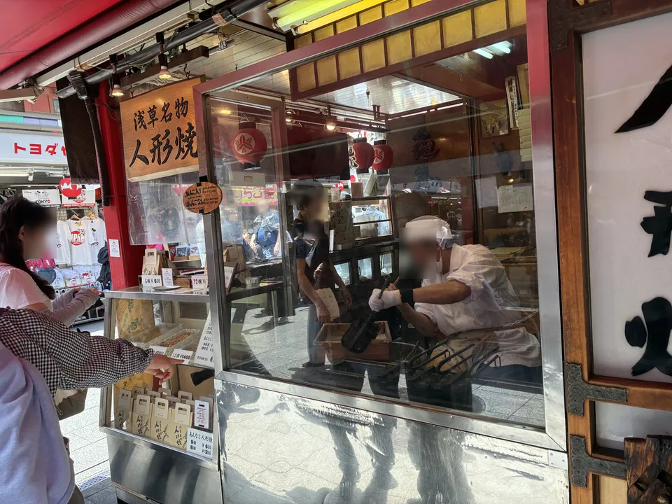 The glass-fronted counter of a 浅草名物 人形焼 (Asakusa specialty ningyo-yaki) shop on Nakamise-dori with a baker in white uniform visible cooking the doll-shaped cake