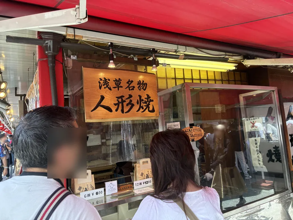 The 浅草名物 人形焼 (Asakusa specialty ningyo-yaki) shop storefront on Nakamise with a large wooden sign above the glass display window and price tags showing 6-piece
