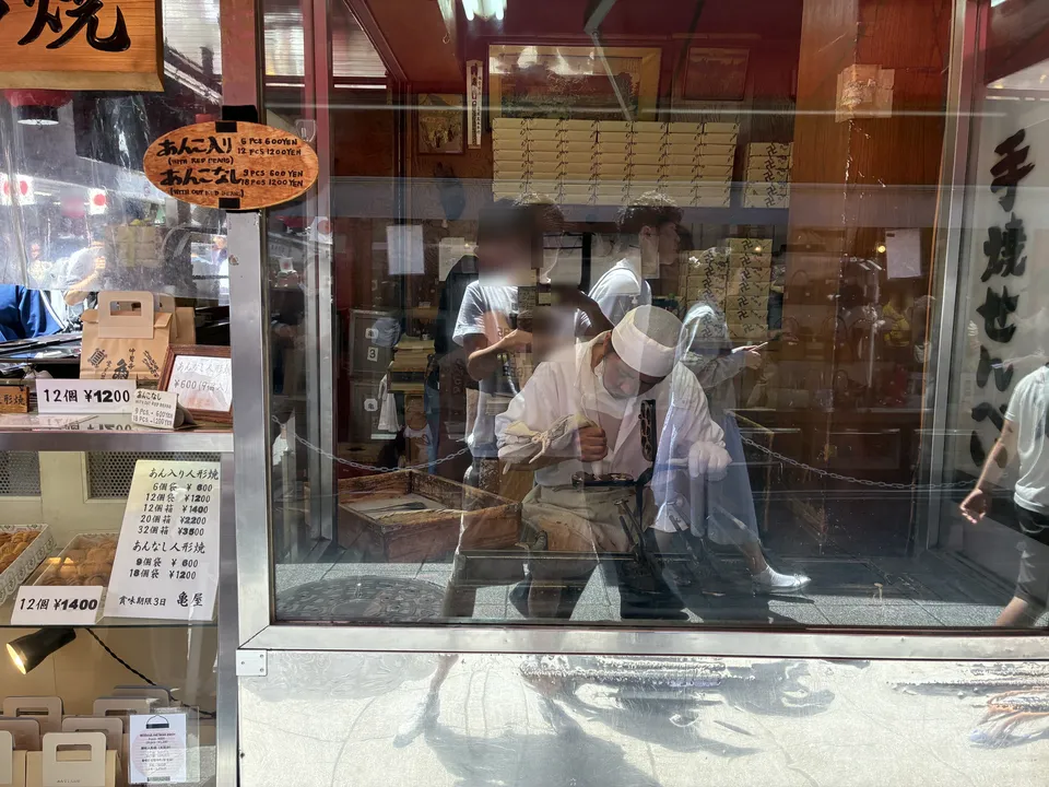Through the glass window of a ningyo-yaki shop on Nakamise, a baker in white uniform fills cast-iron molds, with signs showing あんこ入り (sweet red bean) and あんこなし