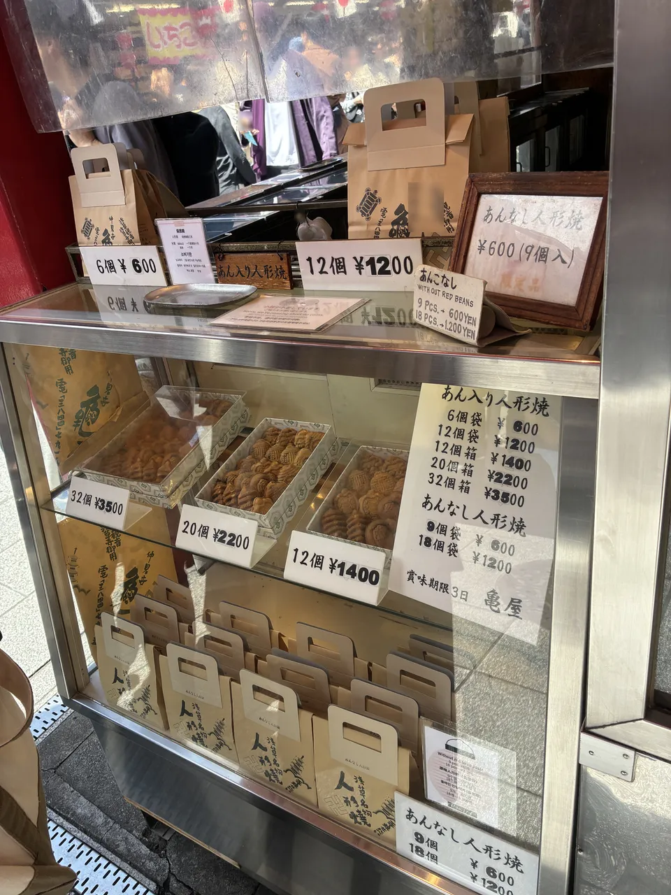 The display case of a ningyo-yaki shop on Nakamise showing packaged boxes of あんこ入り人形焼 (sweet bean doll cakes, 12 pieces ¥1,200) and あんこなし (no bean, 12 pieces ¥1