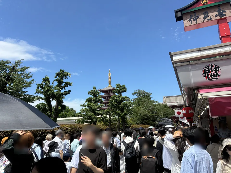 Festival crowds near the end of Nakamise-dori with Senso-ji's five-story pagoda rising behind the trees, a 仲見世 (Nakamise) sign and souvenir shop facade visible 