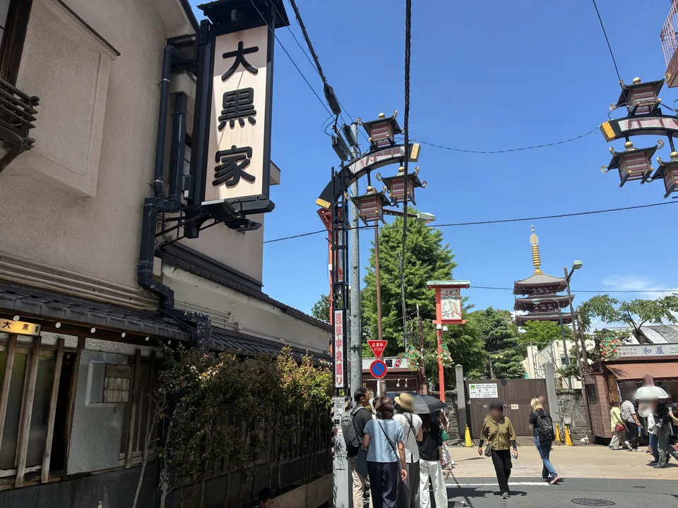 The 大黒家 (Daikoku-ya) tempura restaurant large signboard on the building corner with Senso-ji's five-story pagoda visible above the treetops in the background, a