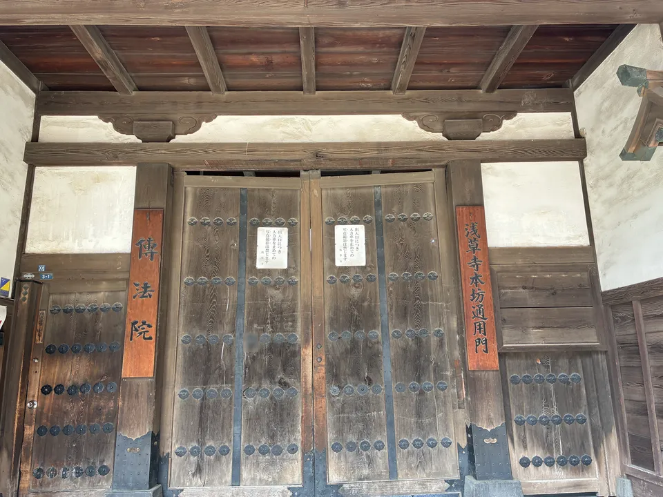 The wooden gate of 伝法院 (Denboin) at Senso-ji showing large nail-studded double doors flanked by orange plaques reading 伝法院 on the left and 浅草寺本坊通用門 (Senso-ji Ho
