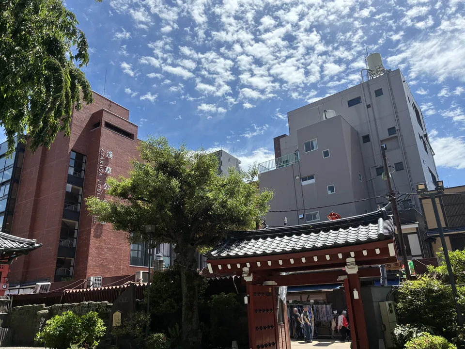 The 浅草公会堂 red brick building rising above tree canopy and a red torii gate at the edge of the Senso-ji precinct in Asakusa, with a gray residential building vis