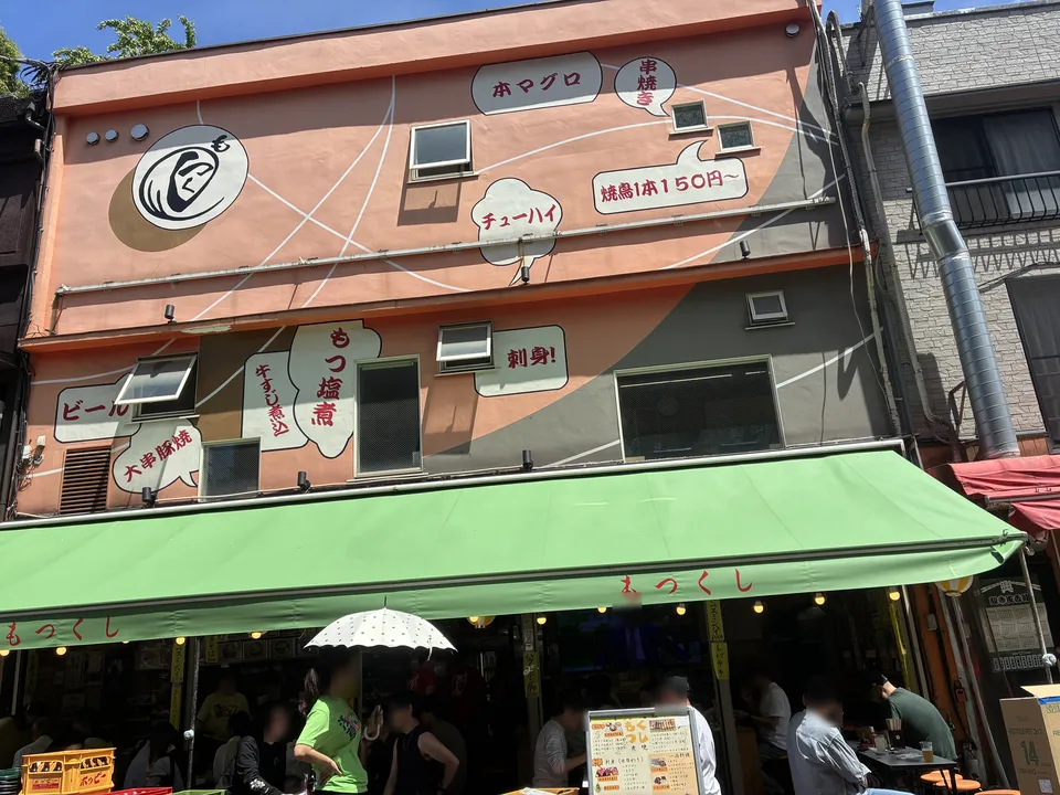 The orange-pink building of もつくし (Motsukushi) restaurant in Asakusa with speech-bubble painted signs listing ホマグロ (bluefin tuna), チューハイ (chuhai), もつ塩煮 (salted o