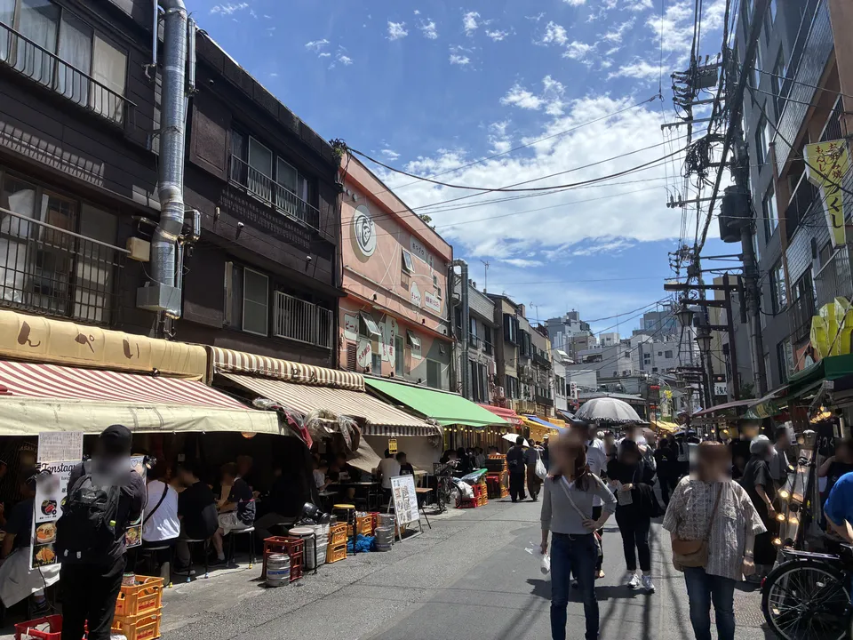 A busy Hoppy Street lane in Asakusa lined with izakaya and food stalls under colorful awnings, with the round-logo Motsukushi building in the background