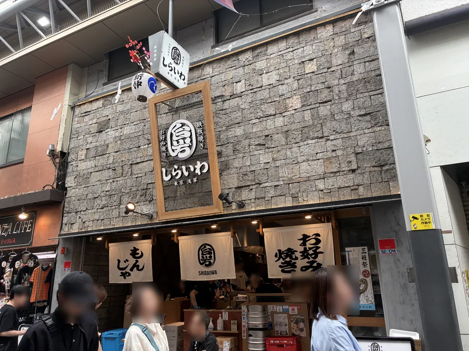 The facade of しらいわ (Shiraiwa) restaurant with a stone-tile exterior, a circular しらいわ logo sign, three white noren curtains reading もんじゃ (monjayaki), SHIRAIWA, a