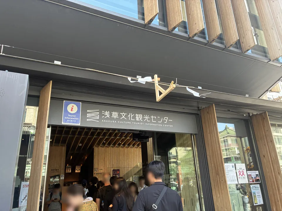 The entrance of 浅草文化観光センター (Asakusa Culture Tourist Information Center) with a modern wooden slatted facade, tourist information ⓘ sign, and visitors at the gla