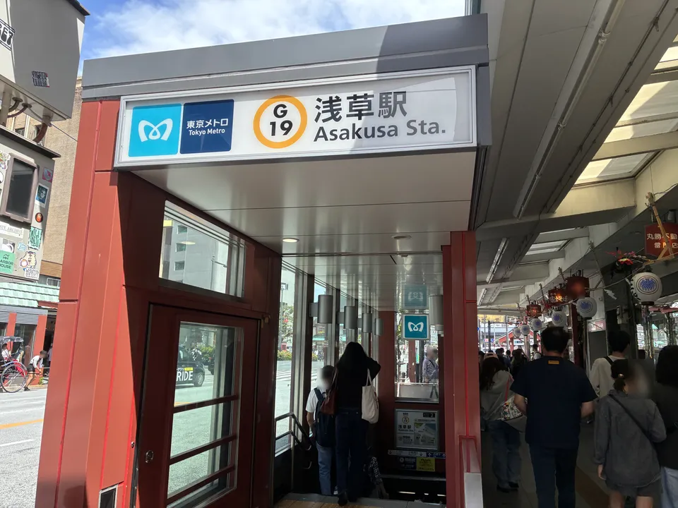Red-framed street entrance of Tokyo Metro Asakusa Station with a white sign reading G19 浅草駅 Asakusa Sta. and people walking down the stairs