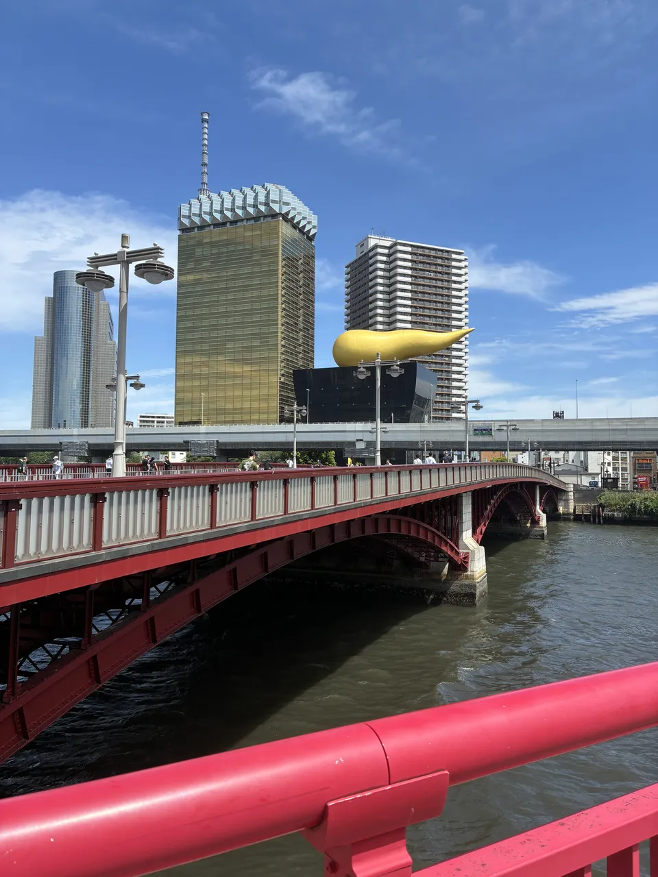 Rotated riverside view from the Azumabashi area showing the red bridge railing, the Sumida River, the gold Flamme d'Or sculpture on the Asahi Beer Hall, and the
