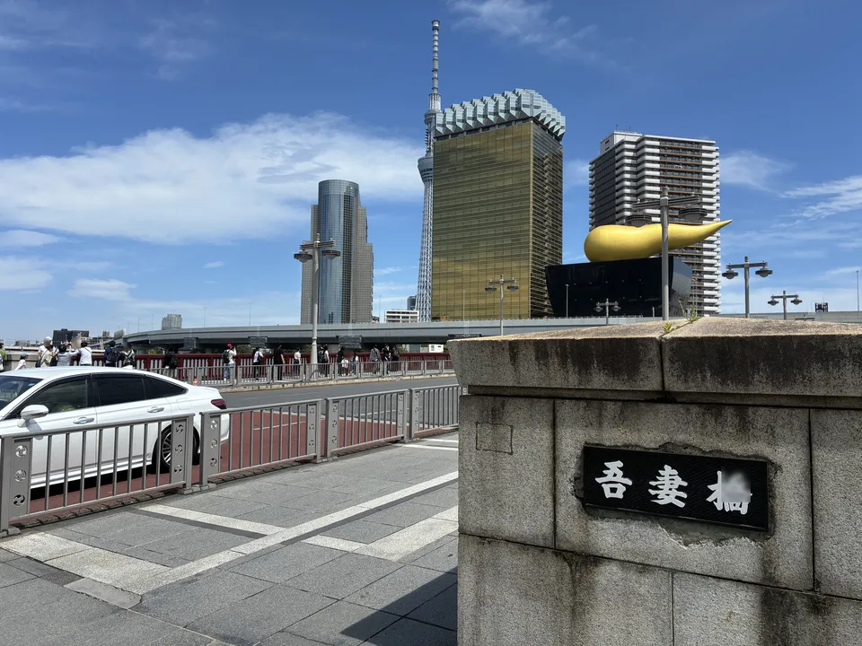 The 吾妻橋 (Azumabashi) nameplate on a concrete bridge abutment with the gold Flamme d'Or sculpture on the Asahi Beer Hall and the Asahi Beer Tower across the Sumi