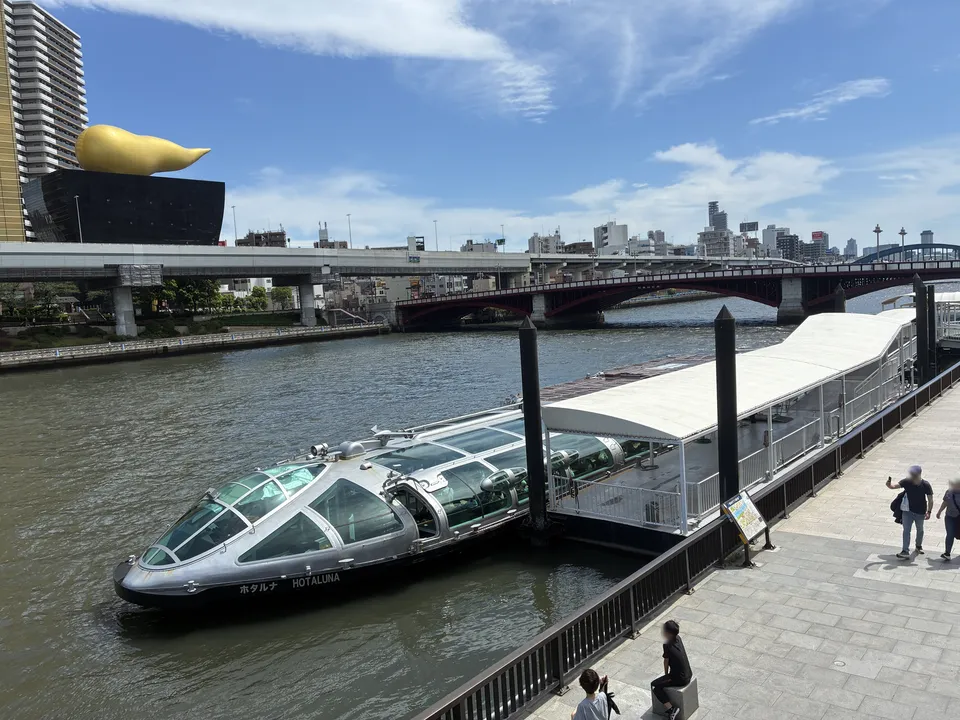 The Tokyo Cruise HOTALUNA futuristic silver water bus docked at the Asakusa Sumida River pier, the gold Flamme d'Or sculpture on the Asahi Beer Hall at upper le