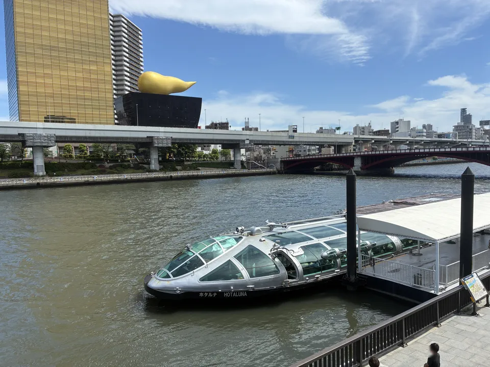 The Tokyo Cruise HOTALUNA silver dome water bus docked at the Asakusa pier with the gold Flamme d'Or sculpture and Asahi Beer Tower behind