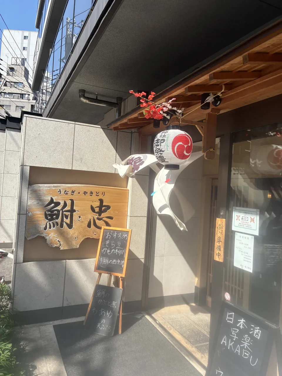 Rotated view of the entrance of Funachu (鮒忠) eel and grilled chicken restaurant with a wooden shop sign board, a round tomoe-pattern paper lantern, artificial p