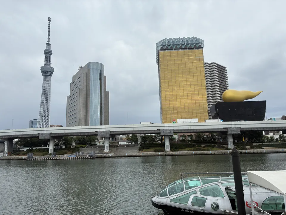 Tokyo Skytree and the gold Asahi Beer building seen across the Sumida River from the Asakusa pier, with the bow of the HIMIKO water bus at the lower right